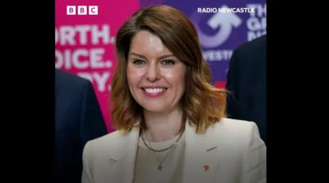 Kim McGuinness is smiling against the backdrop of a BBC studio. She is wearing a white jacket with two gold chains, one a crucifix. She has fair hair and is wearing pink lipstick.
