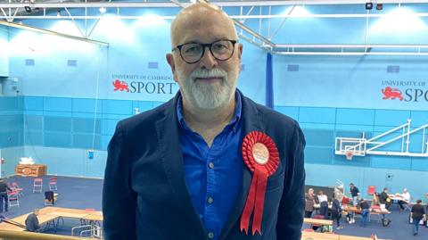 Mike Davey, the Labour leader of Cambridge City Council. He wears a red rosette and the words University of Cambridge Sport is on a wall in the background.