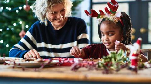 A grandmother helps her granddaughter to decorate Christmas cookies. The girl is wearing reindeer antlers and is picking up decorations. There is a Santa model on the table and a tree behind them.