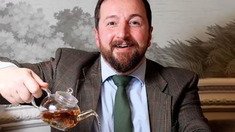 Thomas Robinson wears a suit and pours tea from a glass tea pot into a white cup and saucer. He has short, brown hair and a short, brown beard and is smiling at the camera. 