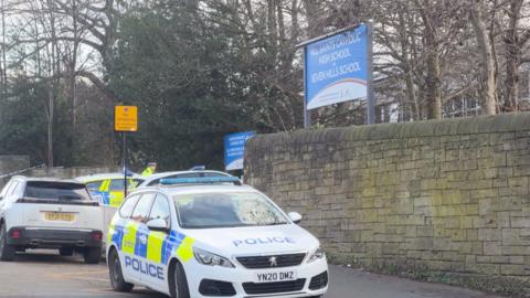 A police car outside a tall stone wall which has a sign for the school on top.