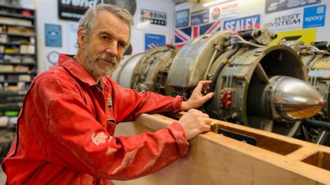 David Aldred has short grey hair and beard and is wearing red overalls. He is stood next  to a jet engine in his garage.