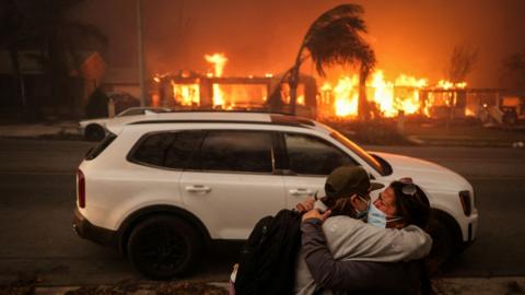 People embrace in front of a car with burning buildings seen in the background as they evacuate following powerful winds fuelling devastating wildfires in the Los Angeles area