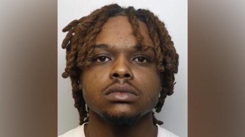 A police custody photograph showing a black man with dreadlocks and a small moustache, staring blankly forward 