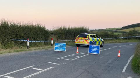 Police car stationary on road which has been cordoned off with blue and white police tape. Blue signs have been put up on the road saying Police Accident and Police Road Closed in English and in Welsh alongside orange and white cones. There is a bend in the road and hills and fields are visible behind.