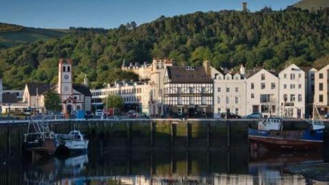 Buildings on a quayside in Ramsey behind boats on the water in the harbour. There are reflections of the buildings on the water and the scenes has a backdrop of hills covered in trees