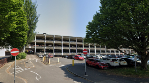 A surface car park full of cars with trees either side of the entrance. Behind it is a grey multi-storey car park with four levels.
