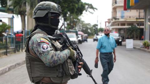 A member of the Ecuadorean Army guards a street in Duran, Ecuador. He is wearing camouflage and a bullet-proof vest, holding a gun, with his finger on the trigger. His face is covered with a black balaclava and dark sunglasses, and he wears a helmet. 