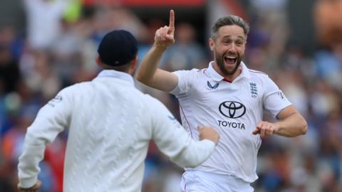 Chris Woakes celebrates a wicket against India by smiling with his finger raised. A team-mate rushes to congratulate him