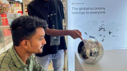 A man in India sitting before an earlier World device, a silver orb, to have his eye scanned as a form of human identification. He has dark hair and a green shirt, and looks straight into the orb, on a desk in front of him, while another man points to it. 