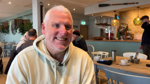 John Kirby smiling, wearing a yellow jumper and sitting in a cafe. There are other Mankind members sitting around a table behind him.