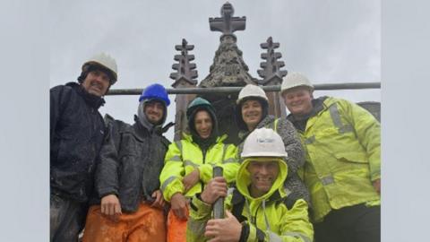 A team of five stonemasons in high visibility protective clothing and helmets smiling at the camera. Parts of Lowther Castle's ornamented pinnacle are visible in the background against a grey sky. 