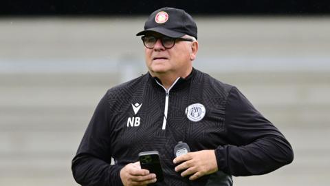 Neil Banfield wearing a black top with a Stevenage badge and NB written on it and a black cap. He is holding a phone in his hand