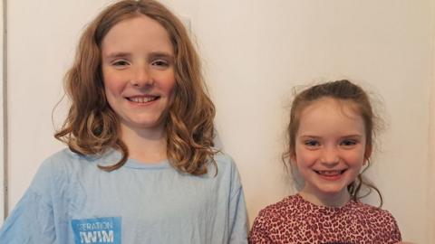 Head and shoulders of Toby and Pippa, standing in front of a cream wall. Toby has long wavy, mousey hair and is wearing a pale blue t-shirt and his sister is standing to his side with her hair tied back and wearing a red and white animal print t-shirt. Both are smiling widely.