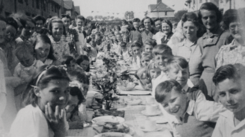 A black and white photo of a street party. There is a long table lined with a tartan cloth. There are white plates and teacups all over the table and in the centre there are flowers going down the line of the table. Lots of children are sat at it, leaning to the camera, surrounded by women holding smaller children.