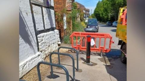 Bike racks are installed next to the black and white walls of the pub. There are still barriers and a road sign up, after the work. Truck wheels are just visible in the road nearby. Cars are parked further away on the village road.