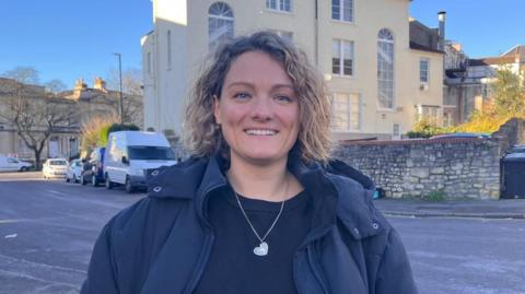 Zoe Armstrong wearing a black coat, looking directly at the camera. She has light brown curly hair and is smiling.