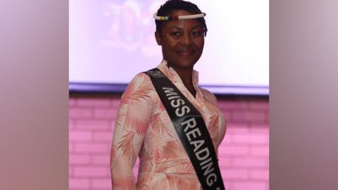 Sarah Sakala is smiling at the camera wearing a pink and white dress, with a white beaded headband and a Miss Reading sash.