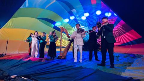 Musicians playing instruments inside a rainbow coloured hot air balloon as it opens up, with lights shining inside it. There are around eight musicians playing violins, a trombone, cello, harp, and clarinet.
The men are wearing white or black smart suits and three of the women are wearing black skirts and turquoise long sleeved silk-style tops and one is in a long sleeved whited top and a long white skirt. They are standing on the rainbow material of the balloon as it surrounds them. 