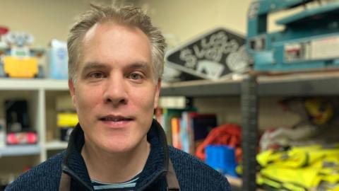 A head and shoulders shot of Alan Dalgairns. He has short grey-brown hair and is wearing a dark and light-blue striped t-shirt with a navy quarter zip jumper. He is also wearing a brown apron. He is smiling at the camera and standing in a room with shelving full of items in the background.