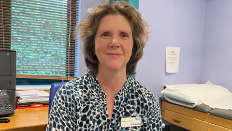 A woman sits in a GP treatment room, behind her is a window and a telephone, to the right of her is a treatment bed. She is smiling, has shoulder length light brown hair and is wearing a turquoise and black patterned blouse with an NHS name tag that reads 'Dr Fleming'.