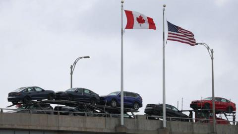 The Canada and US flags flap in the wind on flag polls as a truck stacked with cars for sale drives by