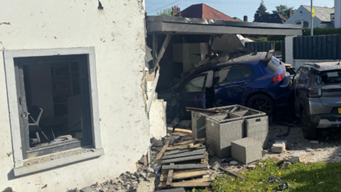 A white house with a navy Volkswagen Golf crashed into it. One wall of the house has been hit and is crumbling.