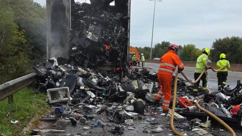 Firefighters battle the flames of a lorry fire on the M5 in Exeter. There is the contents on the lorry on the road surface.