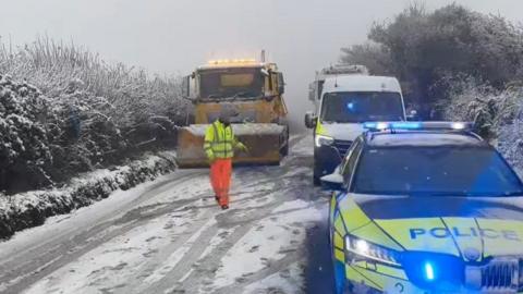 Police car and van on snowy road, worker and snow plough driving towards