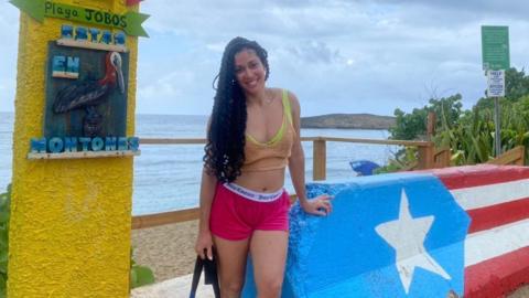 Puerto Rican podcaster Debbie Perez standing beside a concrete barrier painted with the Puerto Rican flag next to a beach