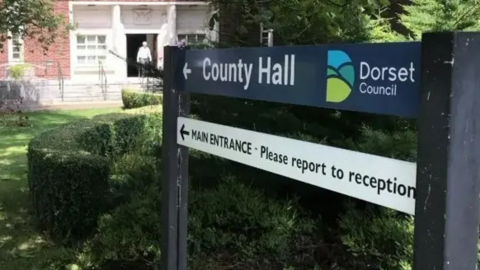 A sign outside Dorset Council's County Hall, which says below "main entrance - please report to reception".