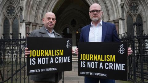 Barry McCaffrey and Trevor Birney outside the Royal Courts of Justice, London, in October. They are holding black signs with yellow and white writing that say 'Journalism is not a crime'. There are black iron gates behind them.