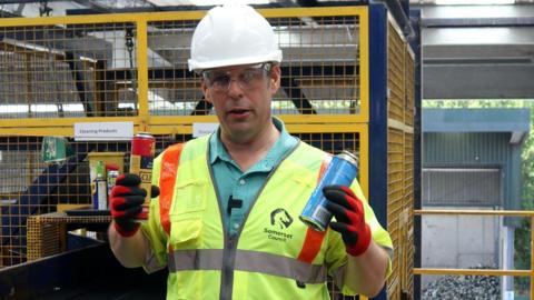A man pictured in a high vis sleeveless jacket, safety goggles and a white hard helmet holding up two gas canisters. One is yellow and red, the other is blue. He is handling them in red gloves. 