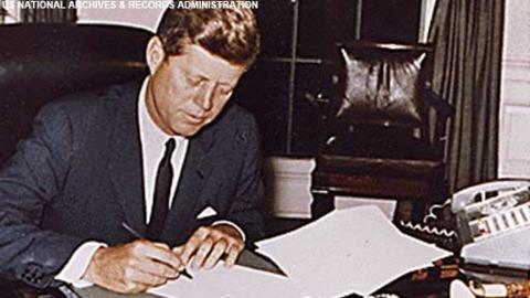 John F Kennedy signing a paper in the Oval Office.