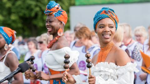 Two women with blue and orange headdresses, white tops and orange necklaces