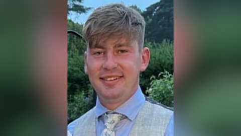 Callum McVeigh. He has dark blonde hair, wearing a light blue shirt, cream waistcoat and cream floral tie.