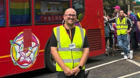 A person in a high‑visibility yellow vest stands in front of a red bus decorated with colorful graphics and the word "PRIDE".