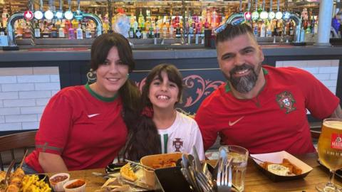 Alexandra, Alice and Sergio Aguiar in a restaurant eating a meal al wearing Portugal football shirts. They are all smiling.