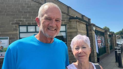 A man in a bright blue t-shirt and short white hair and a stubbly white beard stands next to a woman shorter thanhim with short white hair, she is wearing a white tank top. A brick building is behind them.