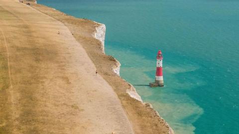 People walking on bone dry grass, along the cliff-top above the lighthouse at Beachy Head, near Eastbourne.