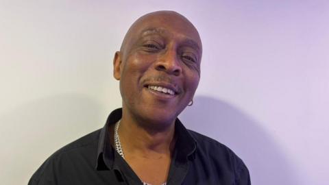 A man wearing a black shirt and silver chain smiles at the camera. 