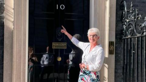 A woman with short white hair, glasses, a white shirt and floral skirt, stands and smiles outside the front door of Number 10 Downing Street while pointing up at the number on the door. 