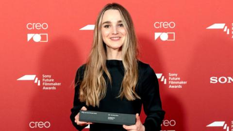 A woman in a black dress with long golden hair stands in front of a red background holding a black oblong box.
