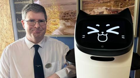 A man wearing a white shirt and navy blue tie with glasses and smiling. He is standing next to a white and black robot with a cat face.