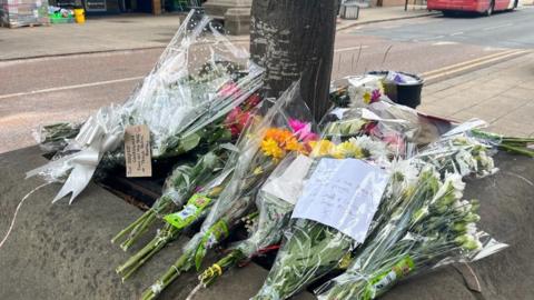 Flowers left around a tree in a street in Batley, West Yorkshire. There are several bunches of flowers, some with notes and ribbons on them.