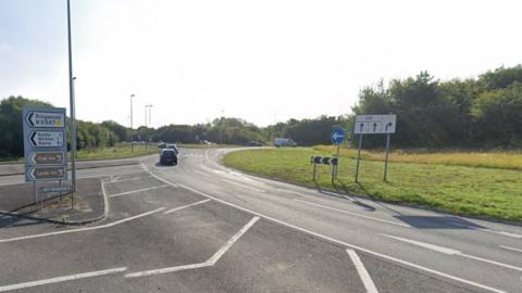 Grassed roundabout with traffic driving around it and various road signs.