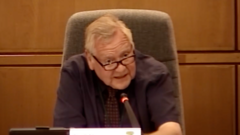 Council leader Mike Gledhill is speaking during a cabinet meeting. He is wearing a brown shirt and tie and is sitting in a brown chair with wooden panelled walls behind.