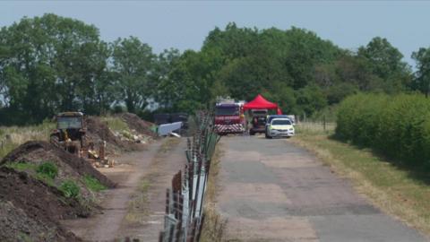 Emergency services parked at the end of a pavement next to fields. There is a fire engine and a police car.