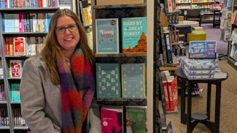 A woman in a grey coat and colourful striped scarf stands next to bookshelves in a shop. She is wearing glasses and has long brown hair parted in the centre. She is smiling.