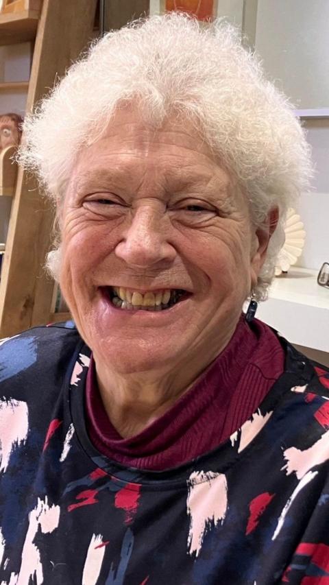 Betty smiles at the camera. She has short white curly hair and wears a navy blue jumper with pink and red plaint splash print all over it.